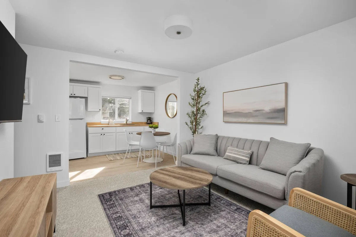 This image showcases a bright and inviting living area featuring a comfortable sofa, a stylish coffee table, a cozy dining nook, and a modern kitchenette, all accented by colorful artwork. The space is designed for relaxation, socializing, and sharing meals, with plenty of natural light creating a warm, welcoming atmosphere.
