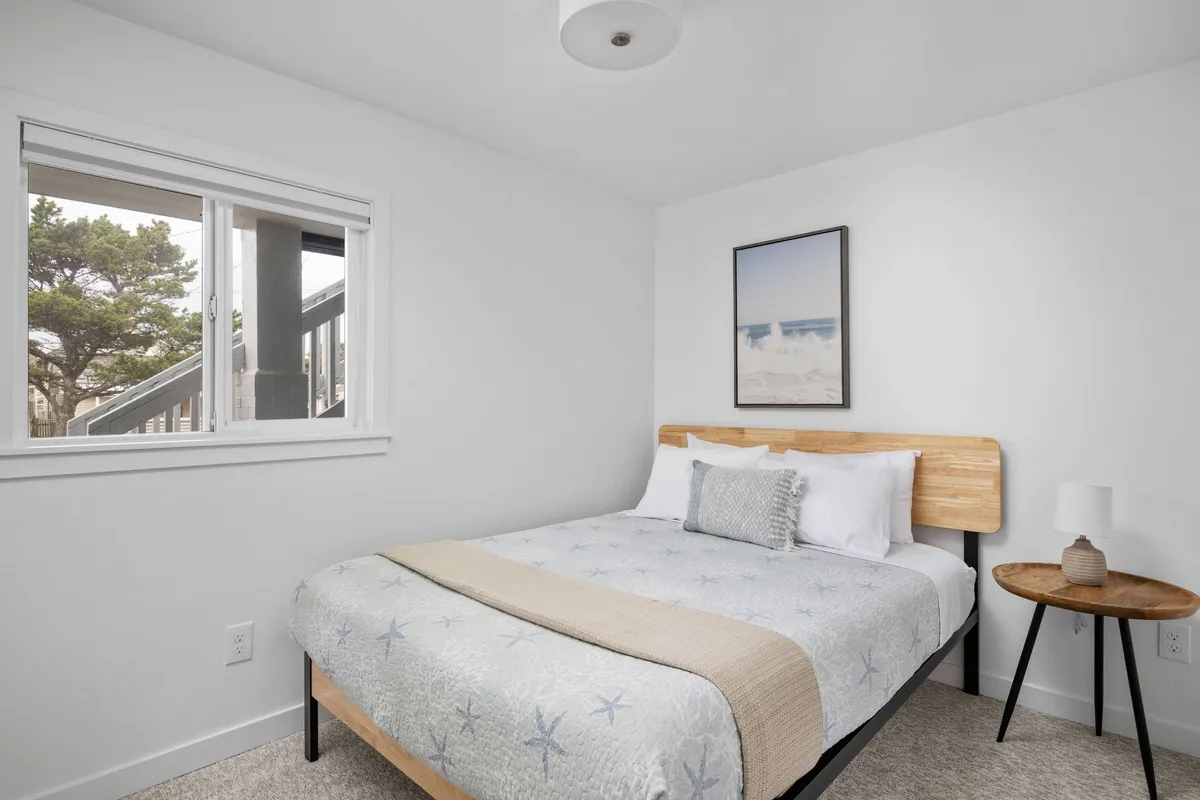 This image presents a tranquil bedroom featuring a plush full bed dressed in crisp linens. Soft lighting lends the room a serene ambiance, while a window allows gentle natural light to filter in. The presence of thoughtfully chosen artwork enhances the sense of retreat, and the overall mood is peaceful and inviting—perfect for unwinding after a day by the beach.