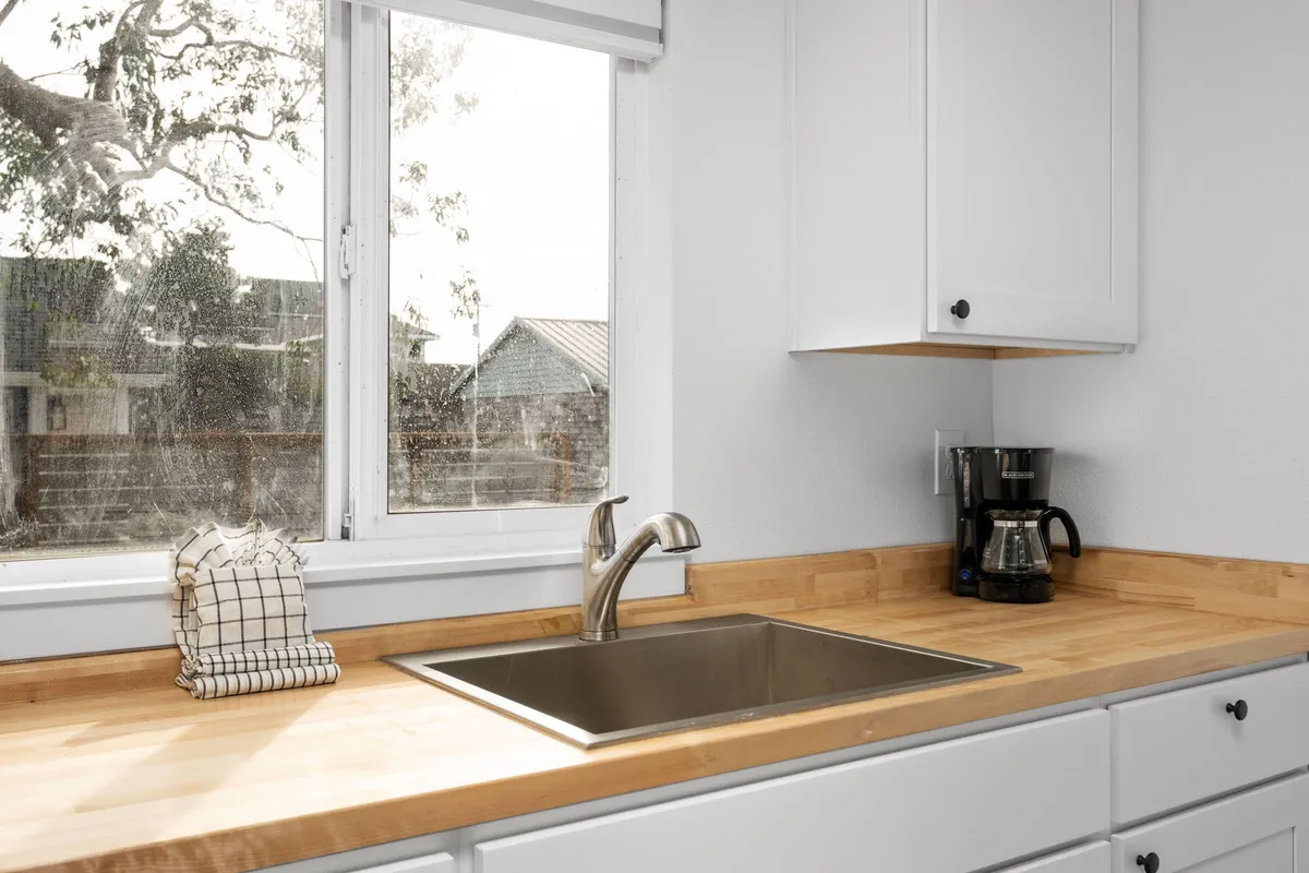 A cozy, communal kitchen space featuring a sink, coffee maker, and clean countertop. The scene suggests guests enjoying morning coffee rituals together, preparing breakfast, and sharing stories while natural light filters in from outside.