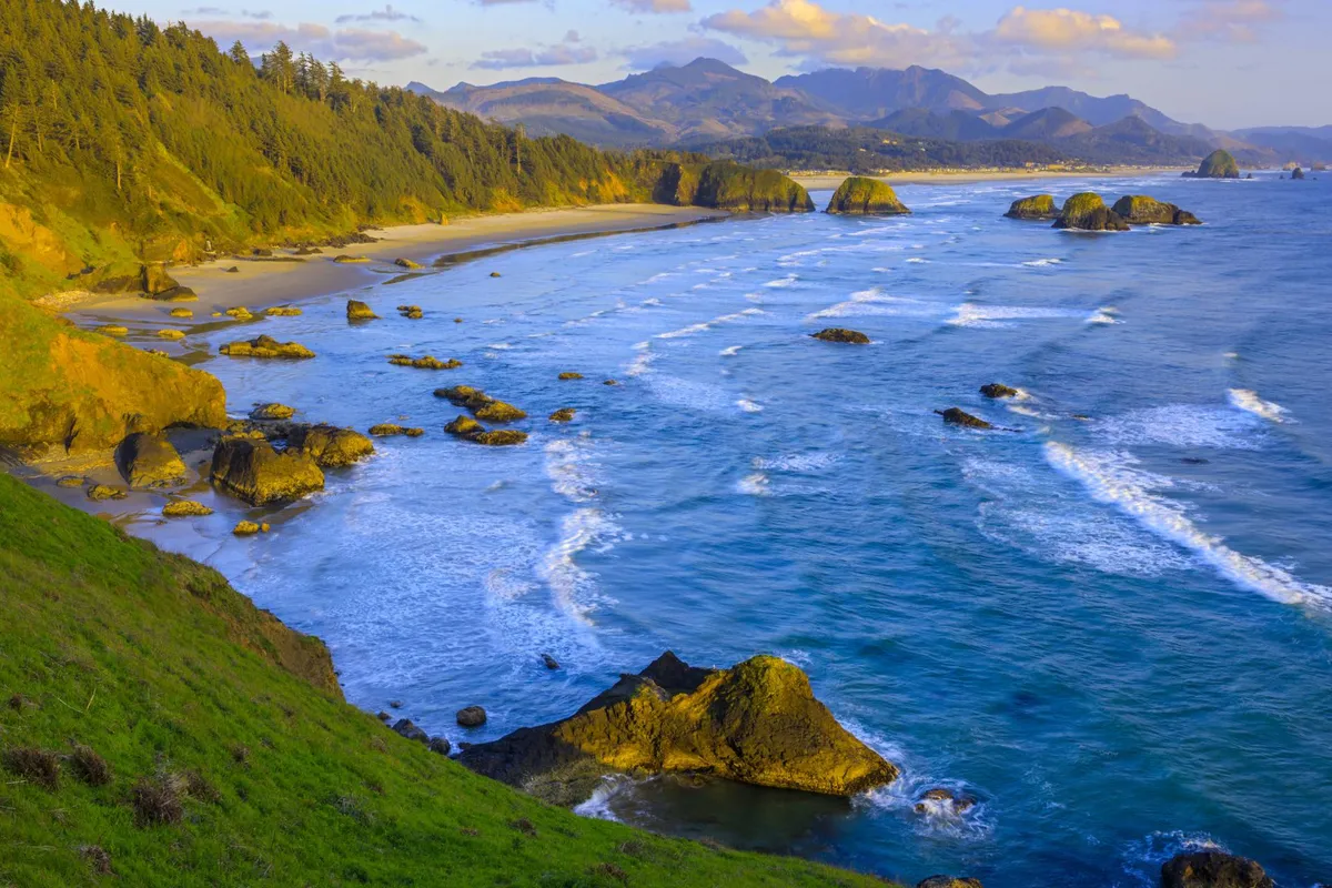 A sweeping view of Rockaway Beach, featuring rolling ocean waves, broad sandy shoreline, dramatic rock formations, distant mountains, and lush tree-covered hills. The expansive vista invites viewers to imagine beachcombing, sunset watching, and enjoying a variety of seaside activities.