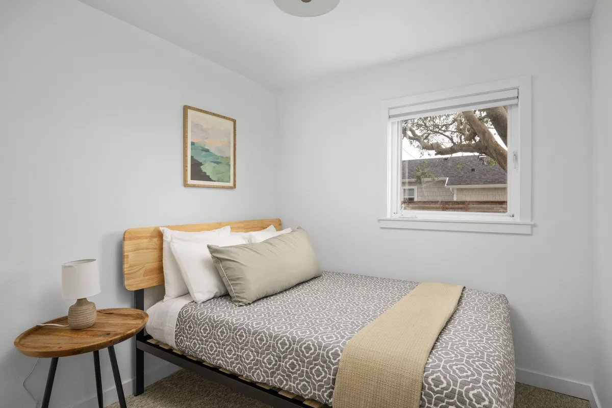 This serene bedroom image features a full bed positioned near a window, with sunlight streaming in to brighten the space. A bedside table and calming artwork add homey touches, while the quiet atmosphere invites guests to rest, read, or simply relax in comfort.